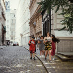 Vier junge Leute gehen an einem sonnigen Tag eine von historischen Gebäuden gesäumte Kopfsteinpflasterstraße entlang. Die Gruppe wirkt entspannt und in ein Gespräch vertieft.