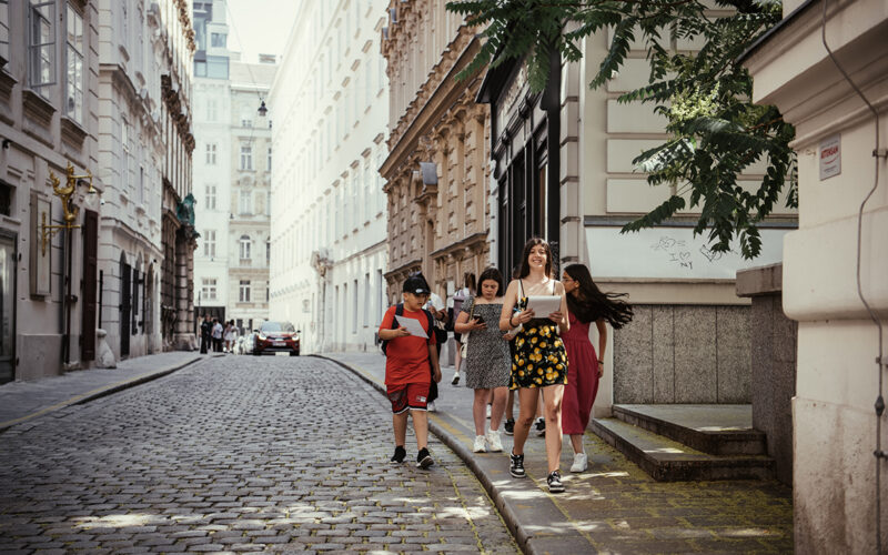 Vier junge Leute gehen an einem sonnigen Tag eine von historischen Gebäuden gesäumte Kopfsteinpflasterstraße entlang. Die Gruppe wirkt entspannt und in ein Gespräch vertieft.
