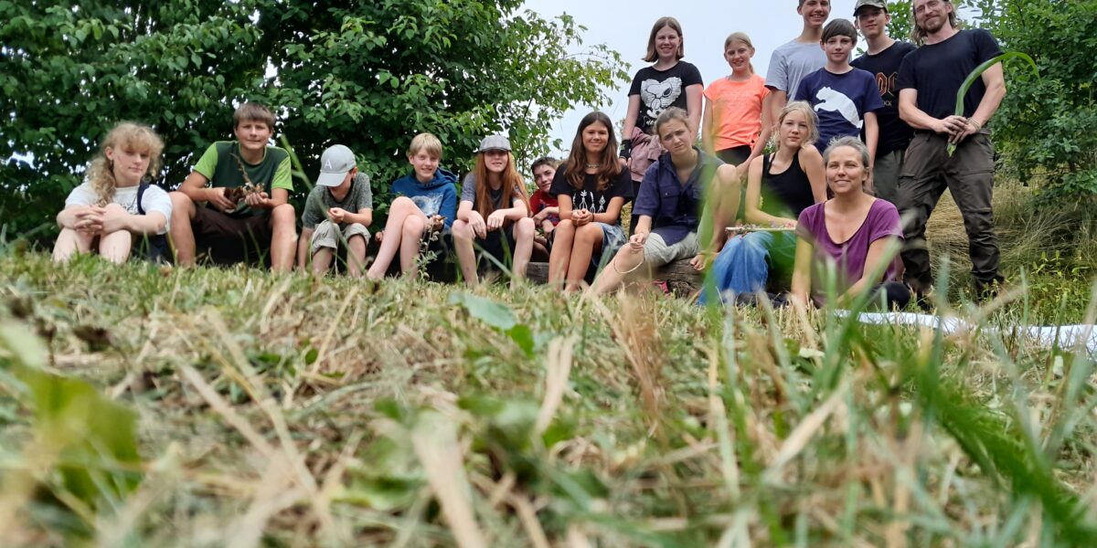 Eine Gruppe von Menschen, darunter Kinder und Erwachsene, sitzt und steht im Freien auf einem grasbewachsenen Hang mit Bäumen im Hintergrund.