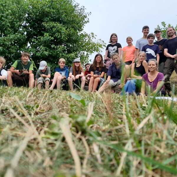 Eine Gruppe von Menschen, darunter Kinder und Erwachsene, sitzt und steht im Freien auf einem grasbewachsenen Hang mit Bäumen im Hintergrund.