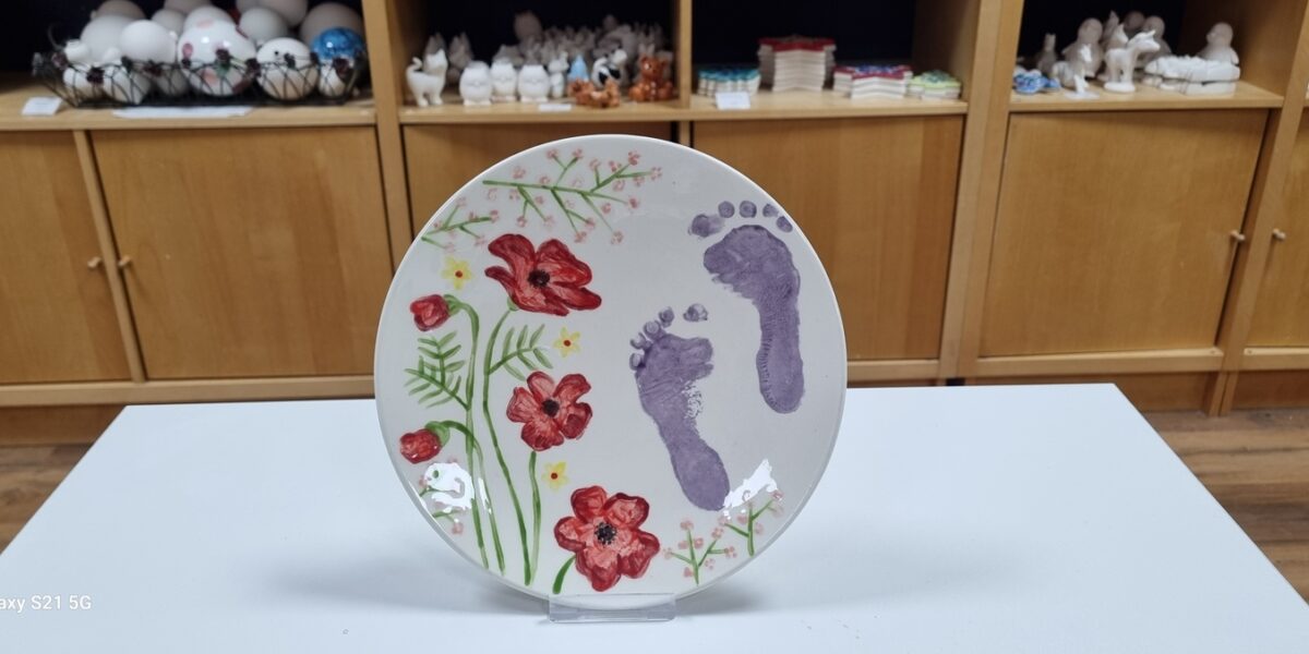 Ein Keramikteller mit gemalten roten Blumen, grünen Blättern und zwei lila Babyfußabdrücken steht aufrecht auf einer weißen Fläche mit Regalen im Hintergrund.
