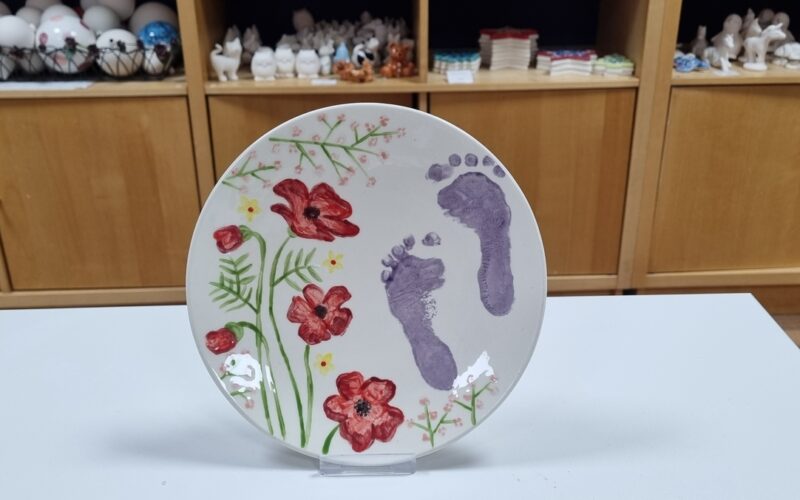 Ein Keramikteller mit gemalten roten Blumen, grünen Blättern und zwei lila Babyfußabdrücken steht aufrecht auf einer weißen Fläche mit Regalen im Hintergrund.