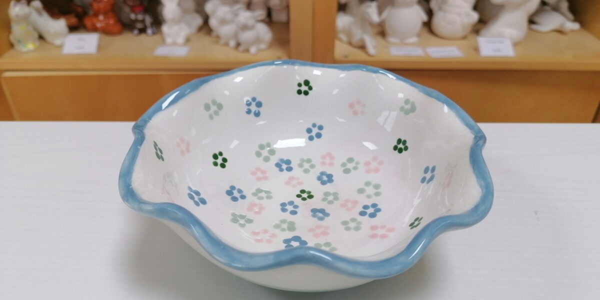 Eine weiße Keramikschale mit gewelltem blauem Rand und gemalten Blumenmustern steht auf einem Tisch; im Hintergrund befinden sich Regale mit unbemalten Keramikfiguren.