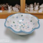Eine weiße Keramikschale mit gewelltem blauem Rand und gemalten Blumenmustern steht auf einem Tisch; im Hintergrund befinden sich Regale mit unbemalten Keramikfiguren.
