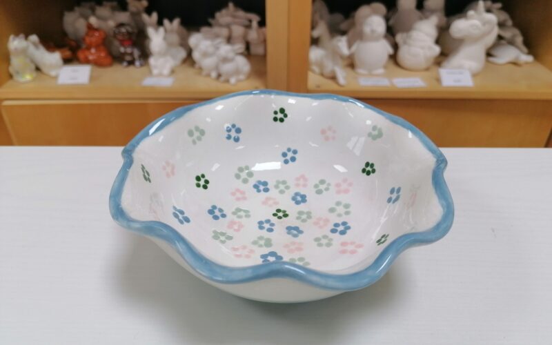 Eine weiße Keramikschale mit gewelltem blauem Rand und gemalten Blumenmustern steht auf einem Tisch; im Hintergrund befinden sich Regale mit unbemalten Keramikfiguren.