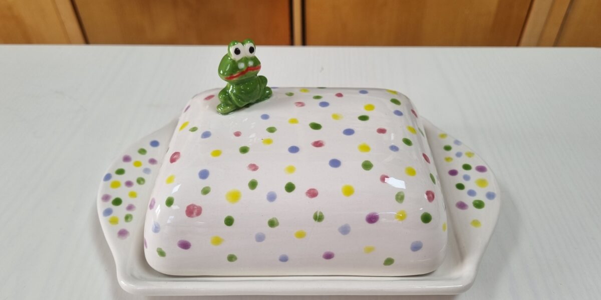 Eine Butterdose aus Keramik mit bunten Tupfen und einer kleinen grünen Froschfigur auf der Spitze, die auf einem weißen Tisch steht.