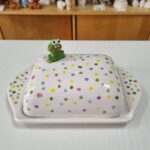 Eine Butterdose aus Keramik mit bunten Tupfen und einer kleinen grünen Froschfigur auf der Spitze, die auf einem weißen Tisch steht.