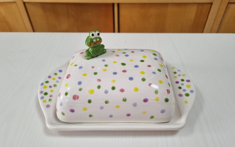 Eine Butterdose aus Keramik mit bunten Tupfen und einer kleinen grünen Froschfigur auf der Spitze, die auf einem weißen Tisch steht.
