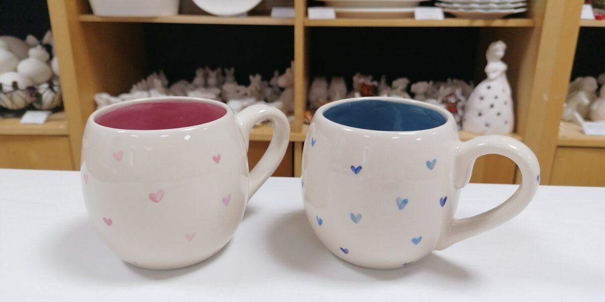 Zwei runde Keramiktassen mit kleinen Herzmustern, eine mit rosafarbenem und eine mit blauem Innenleben, stehen auf einer weißen Fläche vor Regalen mit Tellern und Töpferwaren.