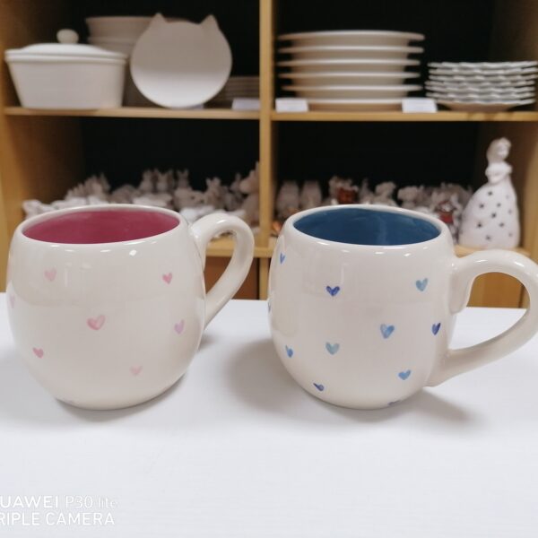 Zwei runde Keramiktassen mit kleinen Herzmustern, eine mit rosafarbenem und eine mit blauem Innenleben, stehen auf einer weißen Fläche vor Regalen mit Tellern und Töpferwaren.