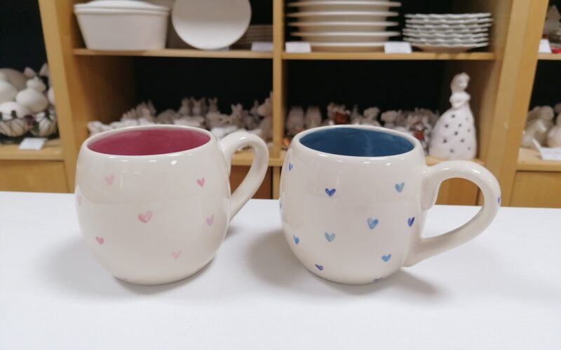 Zwei runde Keramiktassen mit kleinen Herzmustern, eine mit rosafarbenem und eine mit blauem Innenleben, stehen auf einer weißen Fläche vor Regalen mit Tellern und Töpferwaren.