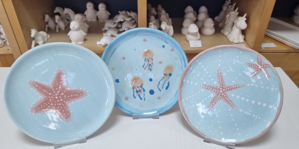 Drei Keramikteller mit Meeresmotiven - Seesterne und Quallen - stehen auf Ständern, während im Hintergrund unbemalte Keramikfiguren auf Regalen stehen.