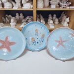 Drei Keramikteller mit Meeresmotiven - Seesterne und Quallen - stehen auf Ständern, während im Hintergrund unbemalte Keramikfiguren auf Regalen stehen.