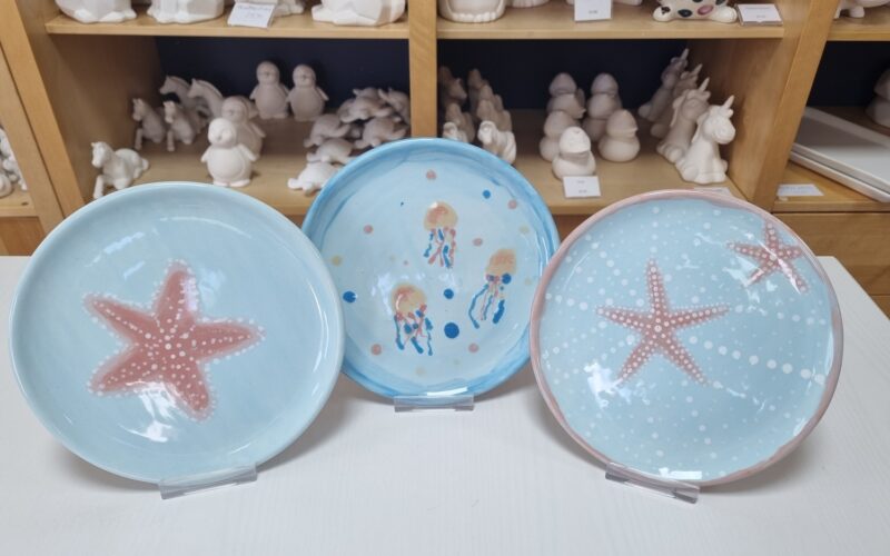 Drei Keramikteller mit Meeresmotiven - Seesterne und Quallen - stehen auf Ständern, während im Hintergrund unbemalte Keramikfiguren auf Regalen stehen.