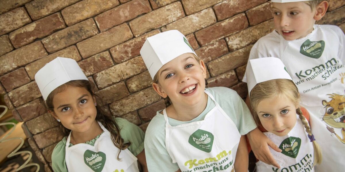 Vier Kinder mit Kochmützen und Schürzen stehen vor einer Backsteinmauer, lächeln und blicken in die Kamera.