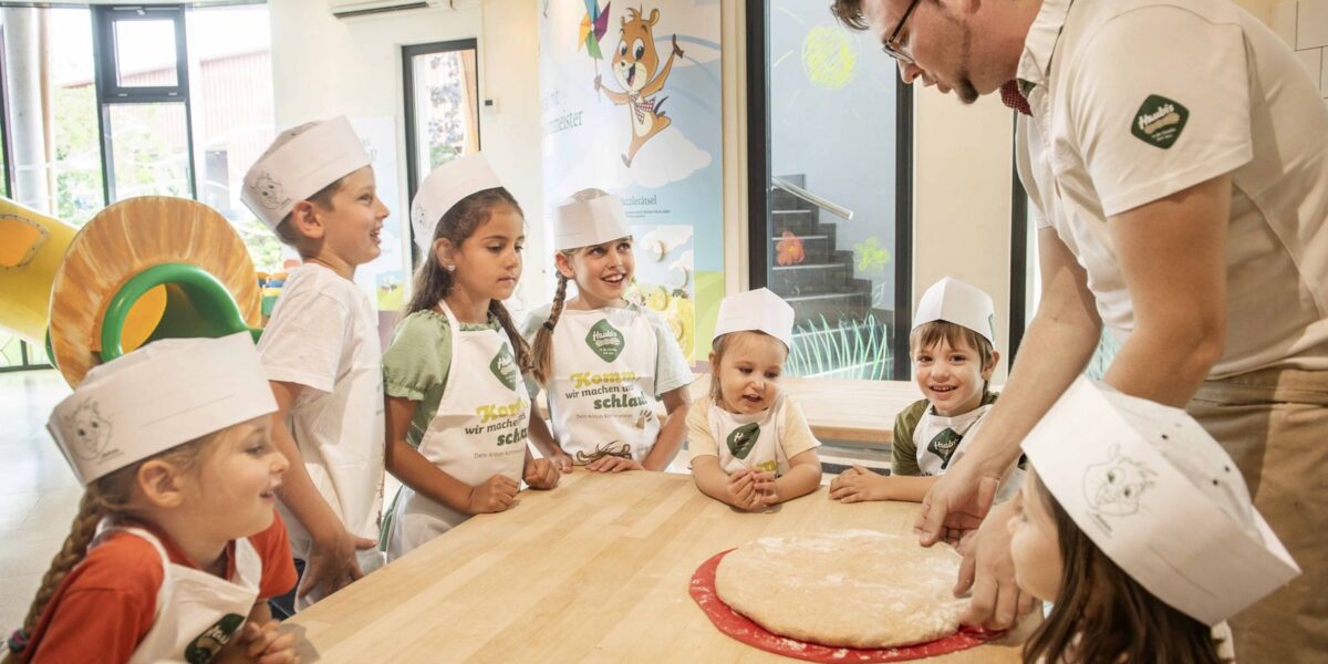 Ein Mann bringt einer Gruppe kleiner Kinder an einem Holztisch in einem hellen Klassenzimmer bei, wie man Teig zubereitet.