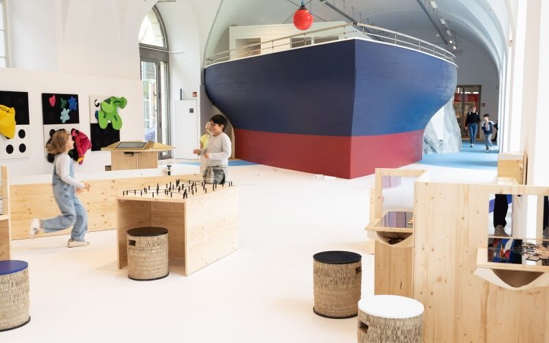 Kinder spielen in einem geräumigen, modernen Raum mit Holzmöbeln und einer großen Bootskonstruktion im Hintergrund.