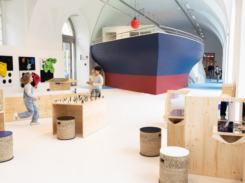 Kinder spielen in einem geräumigen, modernen Raum mit Holzmöbeln und einer großen Bootskonstruktion im Hintergrund.
