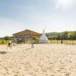 Drei Personen reiten auf einem sandigen Reitplatz in der Nähe eines Holzgebäudes und eines weißen Tipis, mit grünen Bäumen und Hügeln im Hintergrund an einem sonnigen Tag.