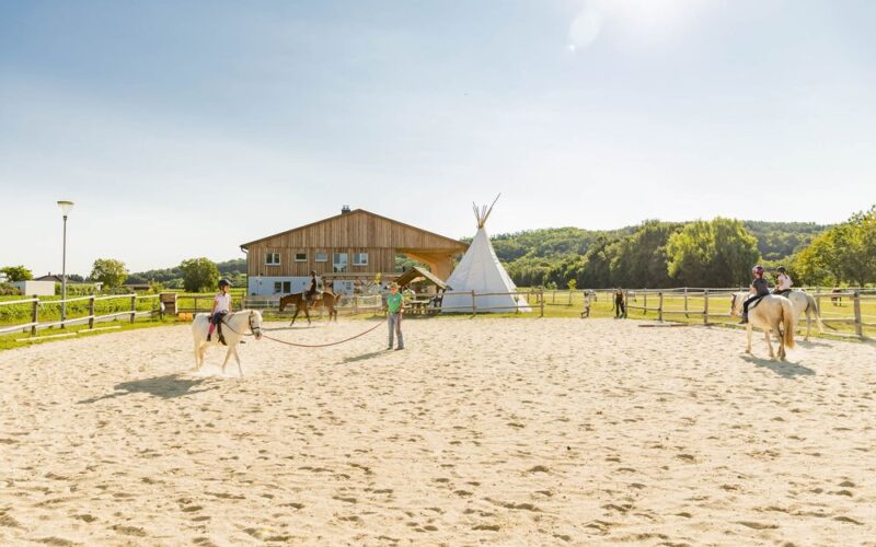 Drei Personen reiten auf einem sandigen Reitplatz in der Nähe eines Holzgebäudes und eines weißen Tipis, mit grünen Bäumen und Hügeln im Hintergrund an einem sonnigen Tag.