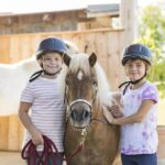 Zwei Kinder mit Helmen stehen neben einem kleinen braunen Pony und lächeln in die Kamera. Im Hintergrund sind ein weißes Pferd und ein Holzzaun zu sehen.