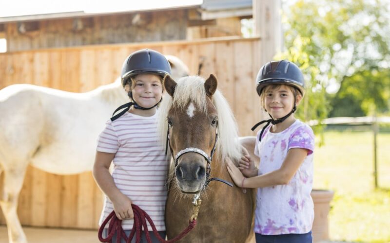Zwei Kinder mit Helmen stehen neben einem kleinen braunen Pony und lächeln in die Kamera. Im Hintergrund sind ein weißes Pferd und ein Holzzaun zu sehen.