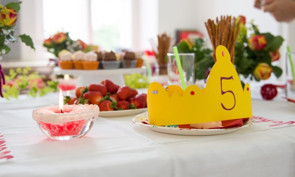 Eine gelbe Papierkrone mit der Zahl 5 steht auf einem Partytisch mit Erdbeeren, Muffins, Blumen und Getränken.