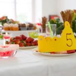 Eine gelbe Papierkrone mit der Zahl 5 steht auf einem Partytisch mit Erdbeeren, Muffins, Blumen und Getränken.