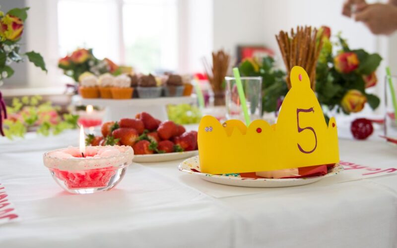 Eine gelbe Papierkrone mit der Zahl 5 steht auf einem Partytisch mit Erdbeeren, Muffins, Blumen und Getränken.