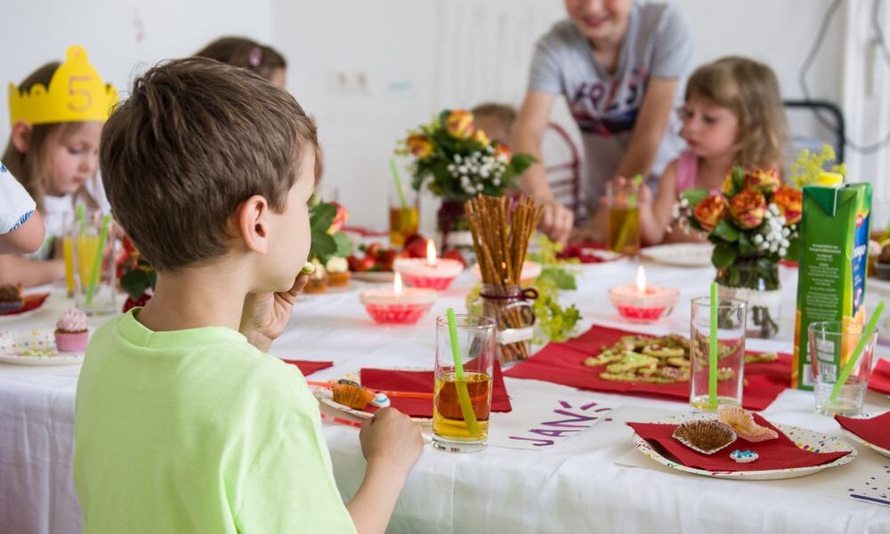 Die Kinder sitzen um einen geschmückten Tisch mit Essen, Getränken, Blumen und Kerzen und scheinen eine Mahlzeit oder eine Feier in einem Raum zu genießen.