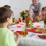 Die Kinder sitzen um einen geschmückten Tisch mit Essen, Getränken, Blumen und Kerzen und scheinen eine Mahlzeit oder eine Feier in einem Raum zu genießen.
