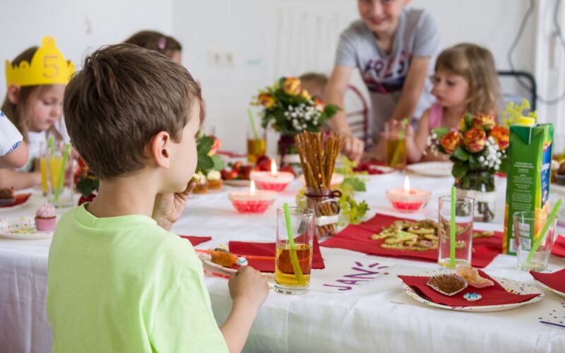 Die Kinder sitzen um einen geschmückten Tisch mit Essen, Getränken, Blumen und Kerzen und scheinen eine Mahlzeit oder eine Feier in einem Raum zu genießen.