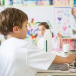 Ein Junge malt an einem Tisch in einem Kunstunterrichtsraum, im Hintergrund sind bunte Kunstwerke und ein anderes Kind zu sehen.