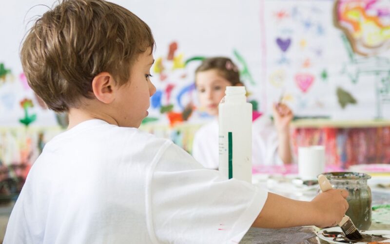 Ein Junge malt an einem Tisch in einem Kunstunterrichtsraum, im Hintergrund sind bunte Kunstwerke und ein anderes Kind zu sehen.