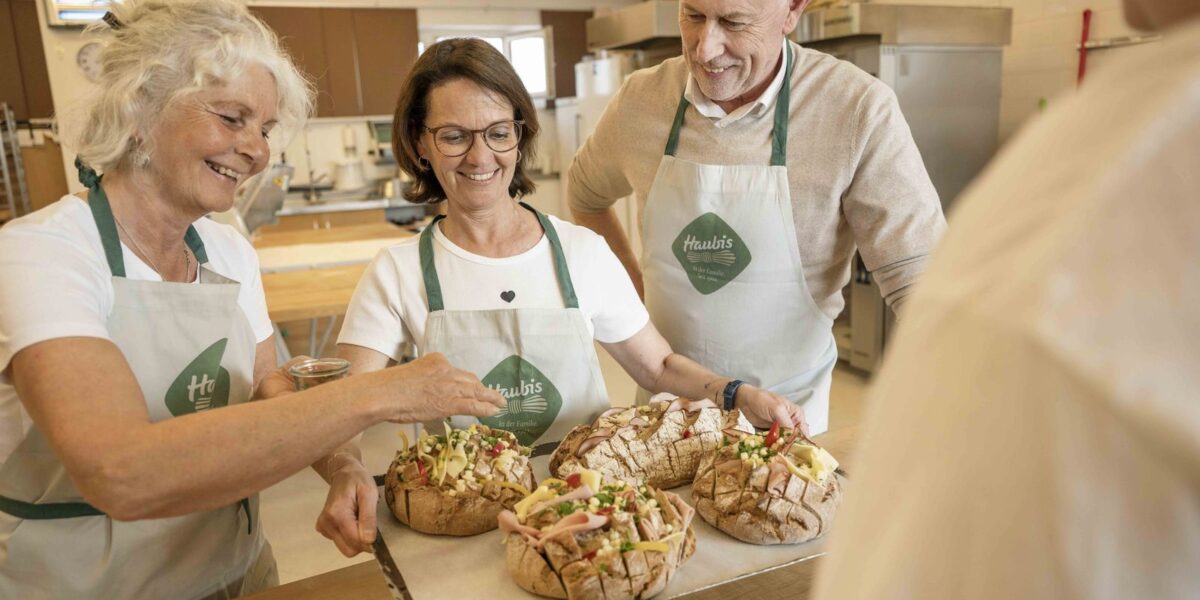 Drei Erwachsene in Schürzen stehen an einer Küchentheke und bereiten lächelnd Brotlaibe zu, die mit verschiedenen Zutaten belegt sind.