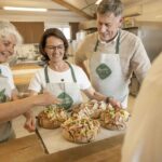 Drei Erwachsene in Schürzen stehen an einer Küchentheke und bereiten lächelnd Brotlaibe zu, die mit verschiedenen Zutaten belegt sind.