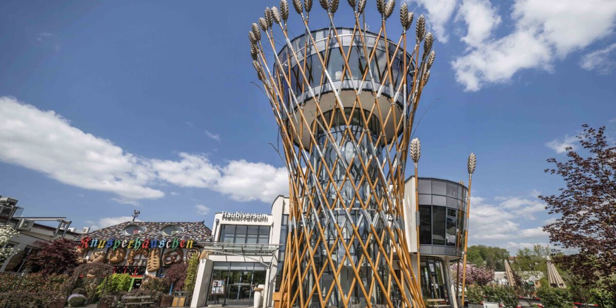 Ein modernes Gebäude mit einem zylindrischen Glasturm, der mit großen Weizenhalmen aus Metall verziert ist, steht unter blauem Himmel in der Nähe eines Restaurants und einer Terrasse.