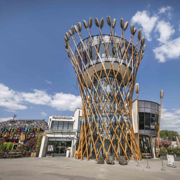 Ein modernes Gebäude mit einem zylindrischen Glasturm, der mit großen Weizenhalmen aus Metall verziert ist, steht unter blauem Himmel in der Nähe eines Restaurants und einer Terrasse.