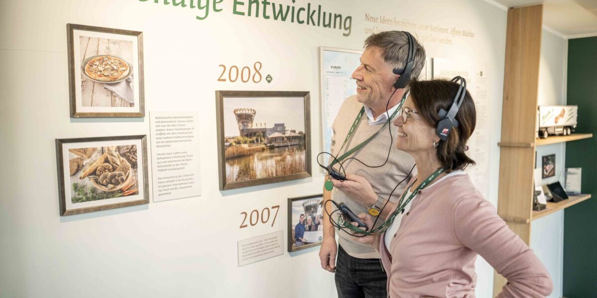 Zwei Erwachsene mit Audioguides betrachten eine Museumsausstellung mit dem Titel „Lebendige Entwicklung“, die Fotos und Texte zu Ereignissen aus den Jahren 2007 und 2008 zeigt.