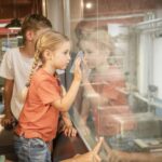 Drei Kinder schauen durch ein Glasfenster; ein Mädchen berührt das Glas, während die anderen etwas auf der anderen Seite beobachten. Innenaufnahme in einer industriellen oder pädagogischen Umgebung.