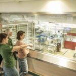 Vier Personen stehen hinter einer Glasscheibe und betrachten eine moderne Industrieanlage, in deren Inneren verschiedene Maschinen und Geräte zu sehen sind.