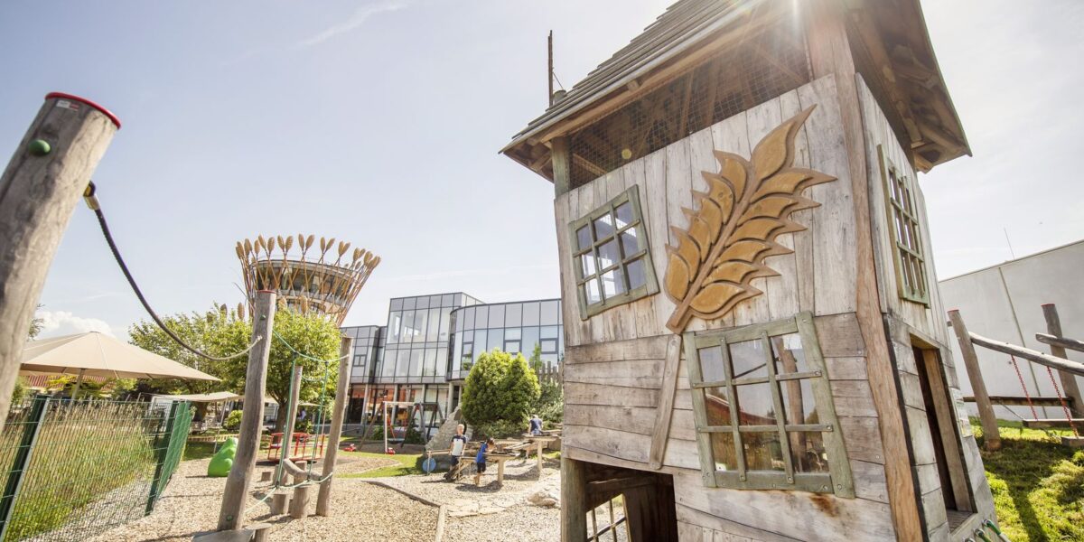 Auf einem Spielplatz steht ein Spielhaus aus Holz mit schrägen Fenstern und einem geschnitzten Weizenmotiv; im Hintergrund sind moderne Glasgebäude und Sitzgelegenheiten im Freien zu sehen.