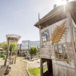 Auf einem Spielplatz steht ein Spielhaus aus Holz mit schrägen Fenstern und einem geschnitzten Weizenmotiv; im Hintergrund sind moderne Glasgebäude und Sitzgelegenheiten im Freien zu sehen.