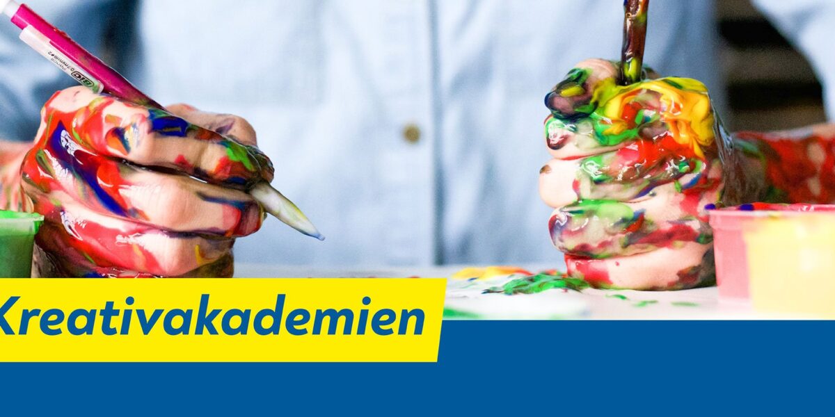 Eine Person mit farbverschmierten Händen hält Pinsel über einen Tisch mit bunten Farben. Auf einem gelb-blauen Banner steht der Text „Kreativakademien“.