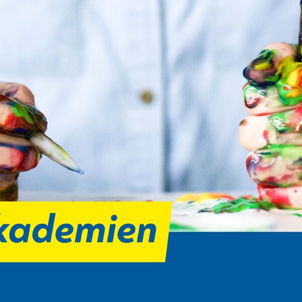 Eine Person mit farbverschmierten Händen hält Pinsel über einen Tisch mit bunten Farben. Auf einem gelb-blauen Banner steht der Text „Kreativakademien“.