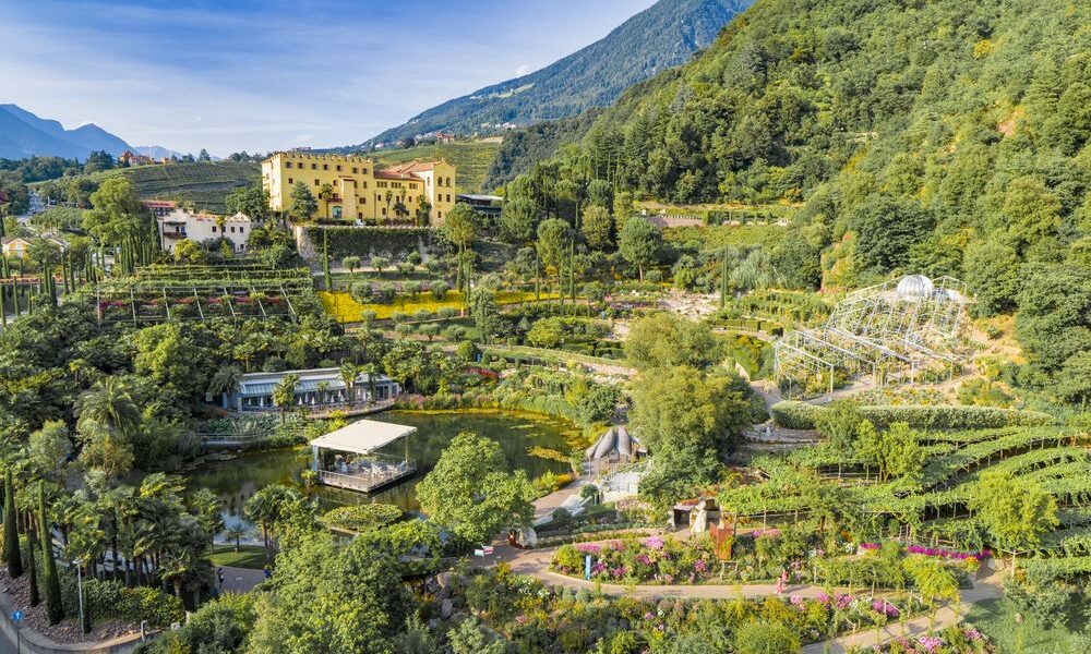 Luftaufnahme einer Gartenanlage mit Spazierwegen, einem Teich und einem großen gelben Gebäude vor bewaldeten Hügeln und Bergen.