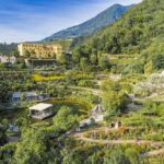 Luftaufnahme einer Gartenanlage mit Spazierwegen, einem Teich und einem großen gelben Gebäude vor bewaldeten Hügeln und Bergen.