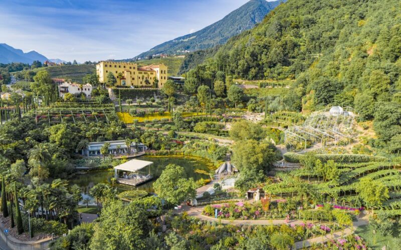 Luftaufnahme einer Gartenanlage mit Spazierwegen, einem Teich und einem großen gelben Gebäude vor bewaldeten Hügeln und Bergen.