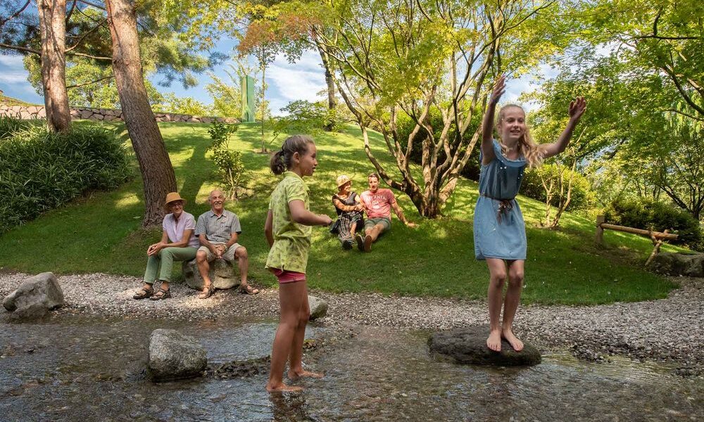 Zwei Mädchen spielen auf Steinen in einem seichten Bach, während vier Erwachsene an einem sonnigen Tag auf der Wiese in der Nähe sitzen, umgeben von Bäumen und Grünpflanzen.