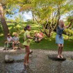 Zwei Mädchen spielen auf Steinen in einem seichten Bach, während vier Erwachsene an einem sonnigen Tag auf der Wiese in der Nähe sitzen, umgeben von Bäumen und Grünpflanzen.